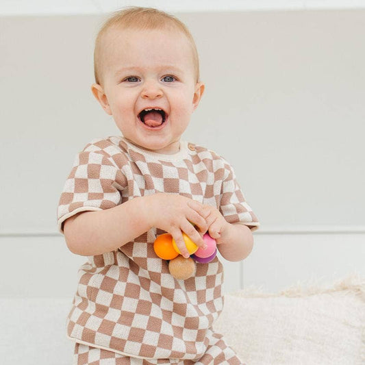 Baby in a checkered outfit holding colorful toys against a plain background