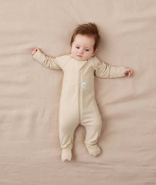 Baby lying on a beige blanket wearing a beige onesie with a small logo on the chest.