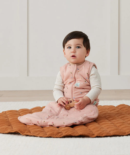 Baby sitting on a brown mat wearing a pink quilted vest and white shirt.