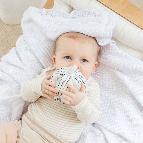 Baby wrapped in a white blanket holding a patterned item, sitting on a light-colored surface.