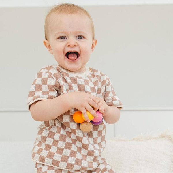 Baby in a checkered outfit holding colorful toys against a plain background