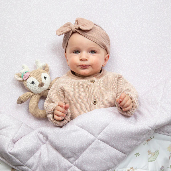 Baby wearing a beige knitted cardigan and headband, holding a plush toy on a white blanket.