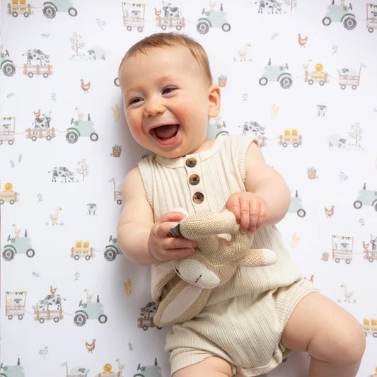 Baby in a beige romper holding a toy, sitting on a patterned surface with vehicles and animals.