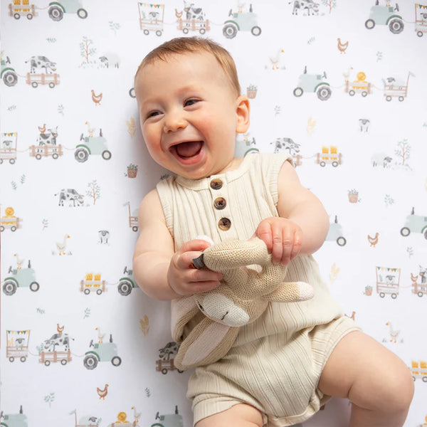 Baby in a beige romper holding a toy, sitting on a patterned surface with vehicles and animals.