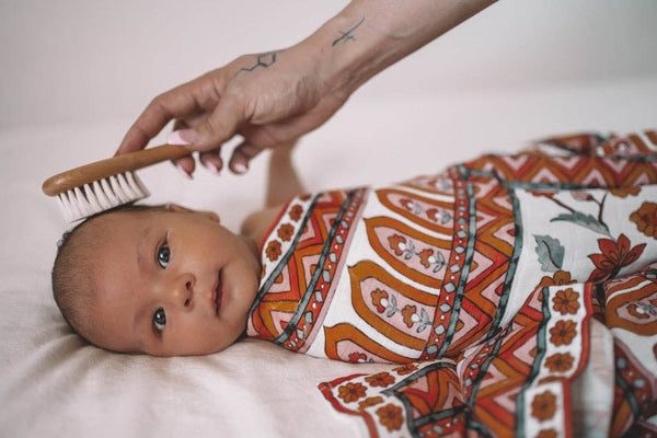 Baby wrapped in a patterned blanket with a hand holding a brush near its head.