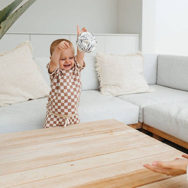 Child in checkered outfit reaching up in a living room with a white couch and wooden coffee table.