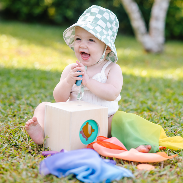 Baby sitting on grass wearing a checkered sun hat and colorful outfit with toys.