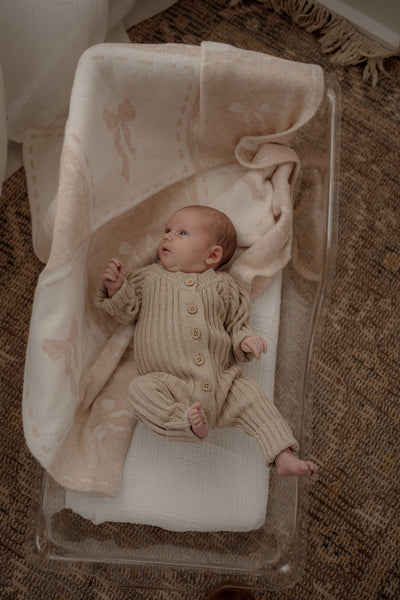 Newborn baby in a beige knitted outfit lying on a white crib with floral patterns.