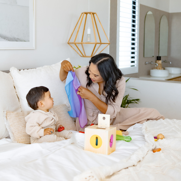 Woman and child playing with toys on a bed in a bright room.