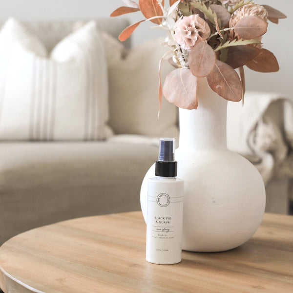 Two bottles of 'Black Fig & Ginger' on a wooden table with a vase of flowers in the background.