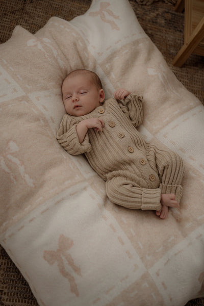 Newborn baby in a beige knitted outfit lying on a patterned blanket.