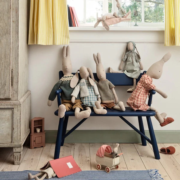 Set of Maileg stuffed animal dolls on a blue bench in a room with yellow curtains and wooden floor.