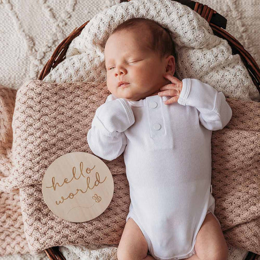 Newborn baby in a white onesie lying on a textured blanket with a 'hello world' sign.