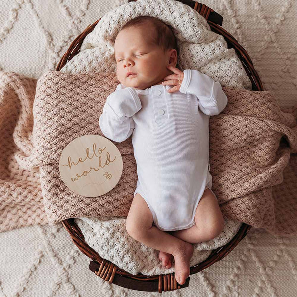 Newborn baby in a white onesie lying in a wicker basket with a 'hello world' sign, surrounded by soft blankets.