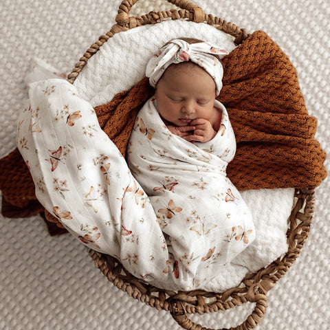 Newborn baby wrapped in a butterfly and floral blanket with a brown blanket, lying in a wicker basket on a textured surface.