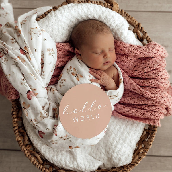 Newborn baby wrapped in a floral blanket with 'hello world' card in a wicker basket.