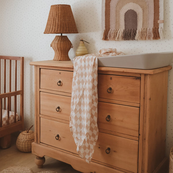 Wooden dresser with a lamp and towel in a room with a crib and wall art.