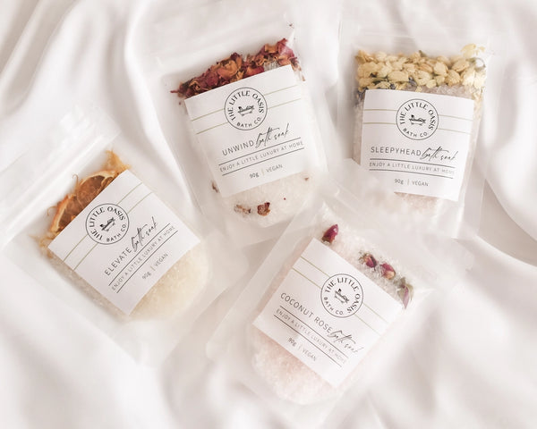 Four packages of 'Little Oasis Bath Co ' bath salts on a white fabric background