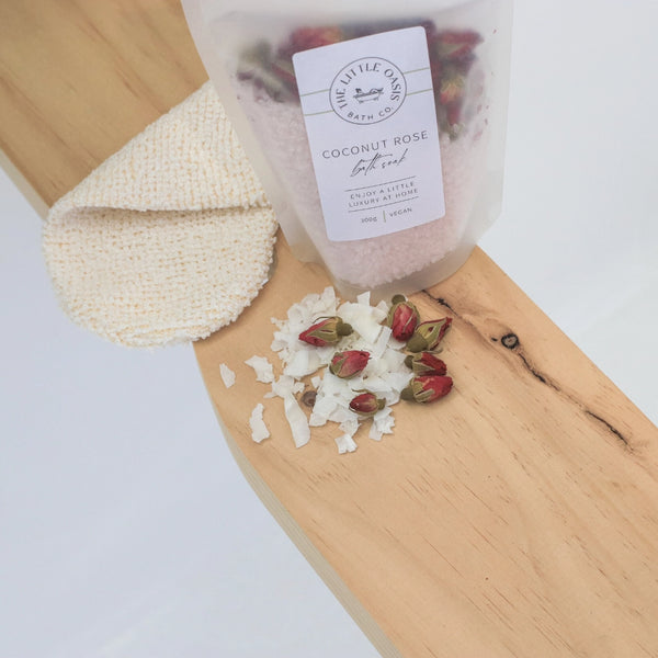 Coconut rose bath salt with a loofah and rose petals on a wooden board