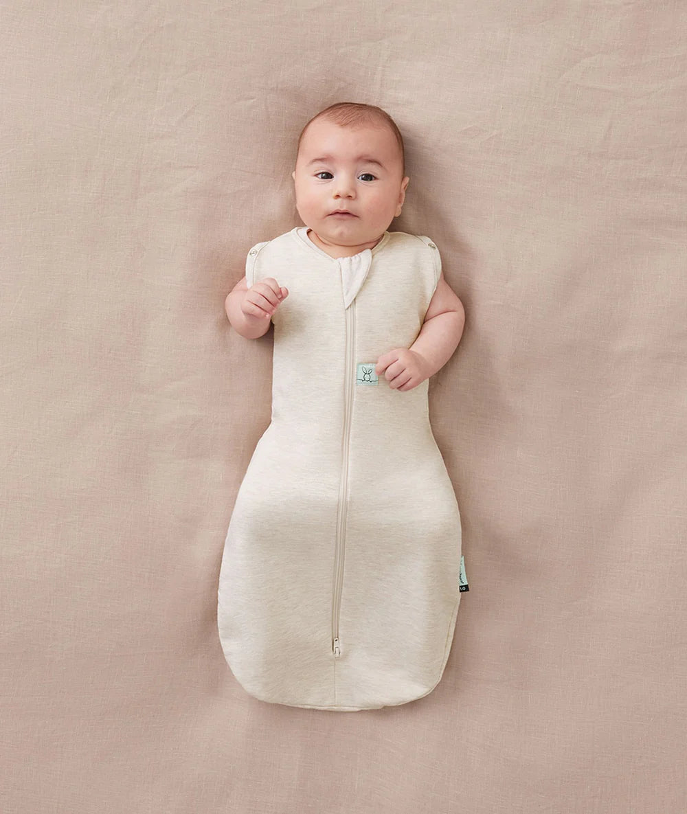 Baby lying on a beige surface wearing a white sleep bag with a small green tag.