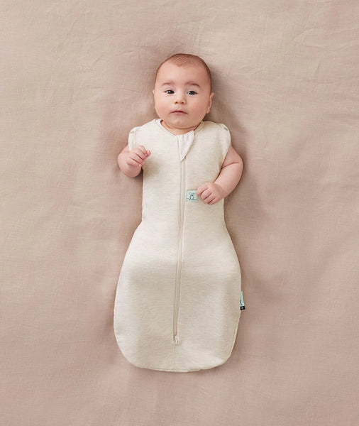 Baby lying on a beige surface wearing a white sleep bag with a small green tag.