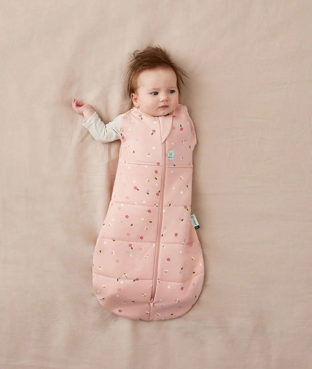 Baby lying on a beige blanket wearing a pink sleeping bag with a pattern.