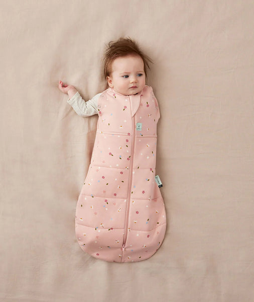 Baby lying on a beige blanket wearing a pink sleeping bag with a pattern.