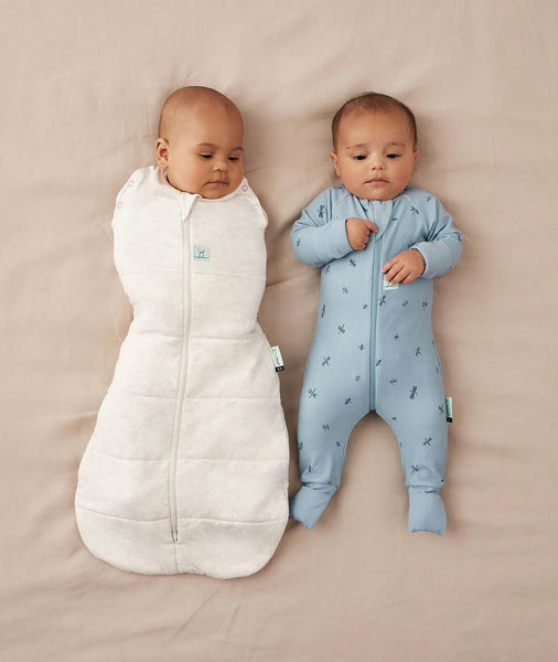 Two babies, one in a white sleeping bag and the other in a blue onesie, lying on a beige surface.