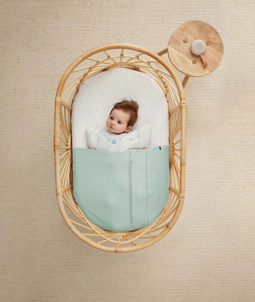 Baby in a rattan crib with a light green mattress against a beige wall.