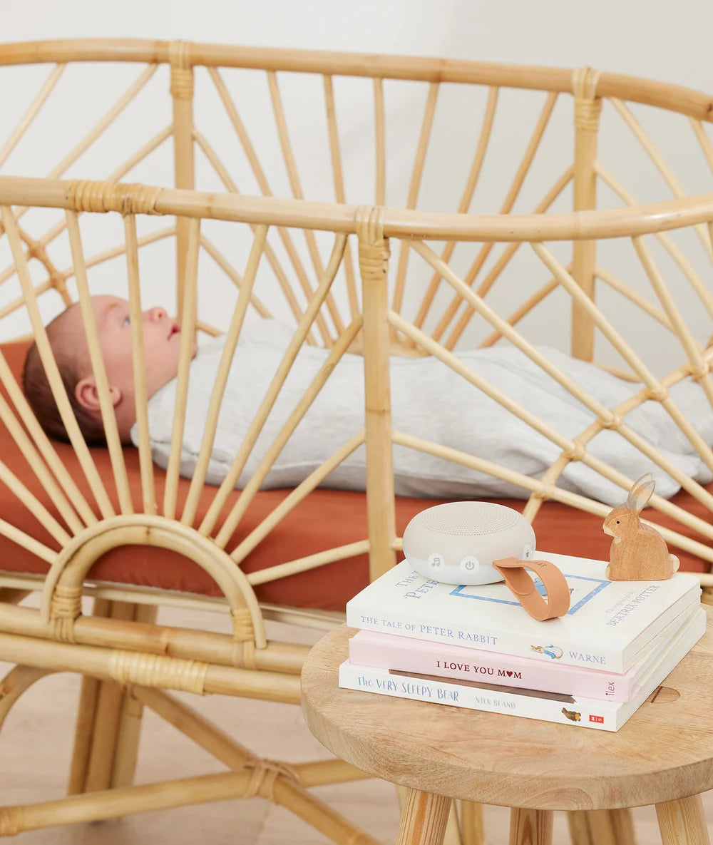 Baby lying in a rattan crib with a small table next to it holding books and a toy.