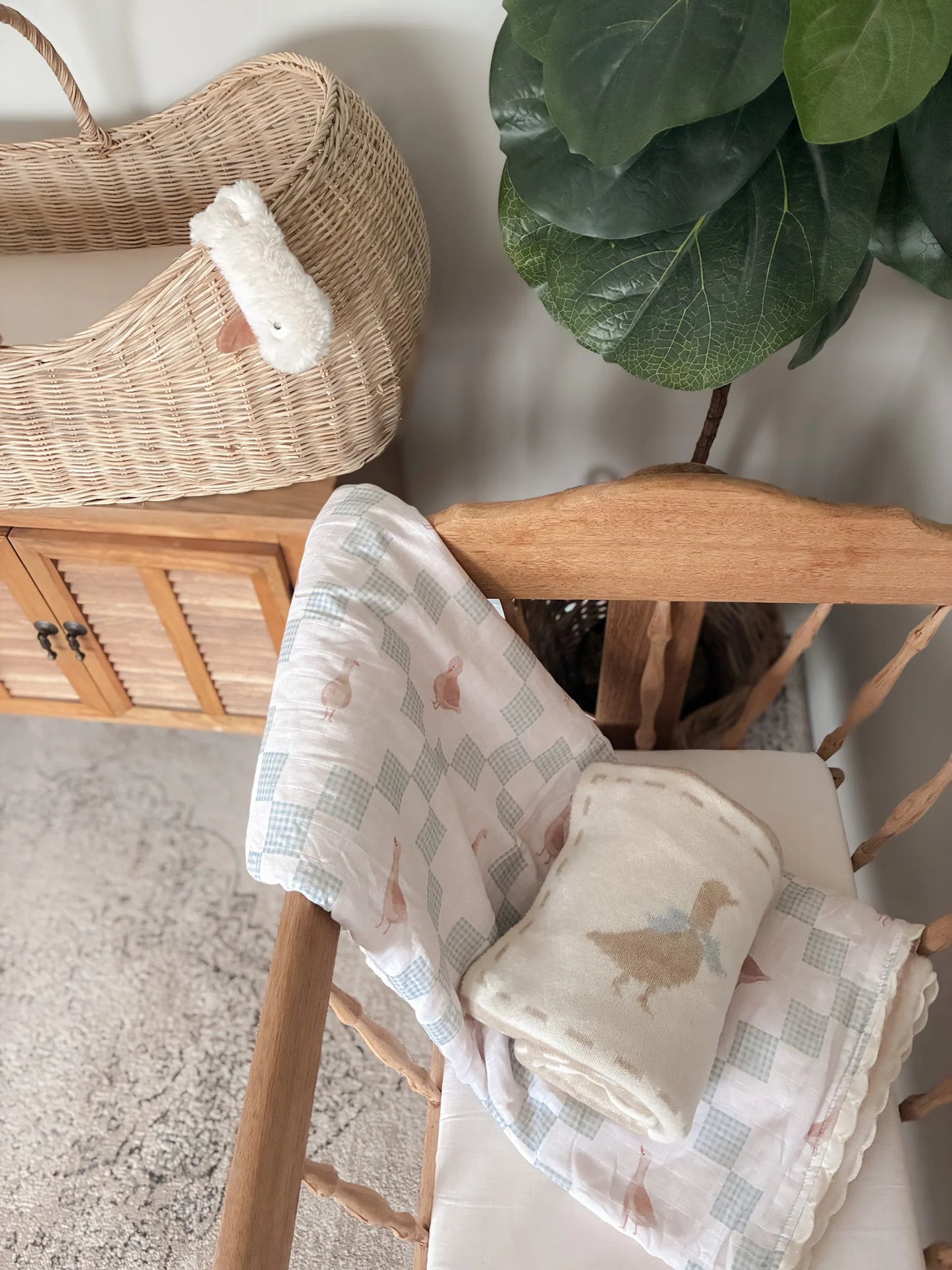 Baby blankets with duck designs on a wooden chair, with a wicker basket and plant in the background.