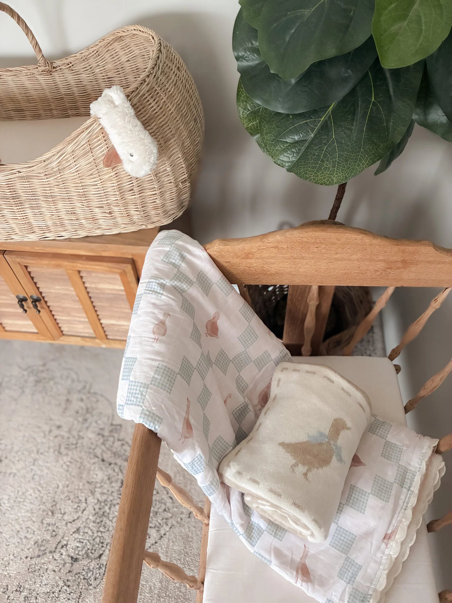 Baby blankets with duck designs on a wooden chair, with a wicker basket and plant in the background.