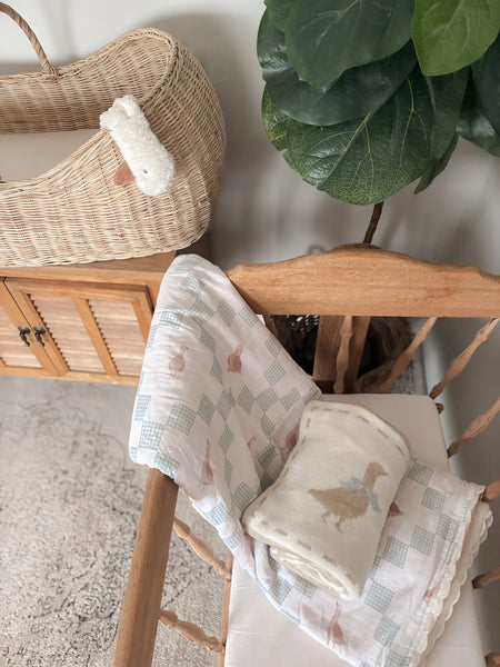 Baby blankets with duck designs on a wooden chair, with a wicker basket and plant in the background.