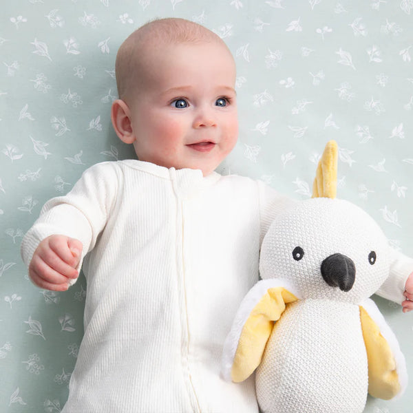 Baby holding a white and yellow stuffed parrot toy against a light blue floral-patterned wall.