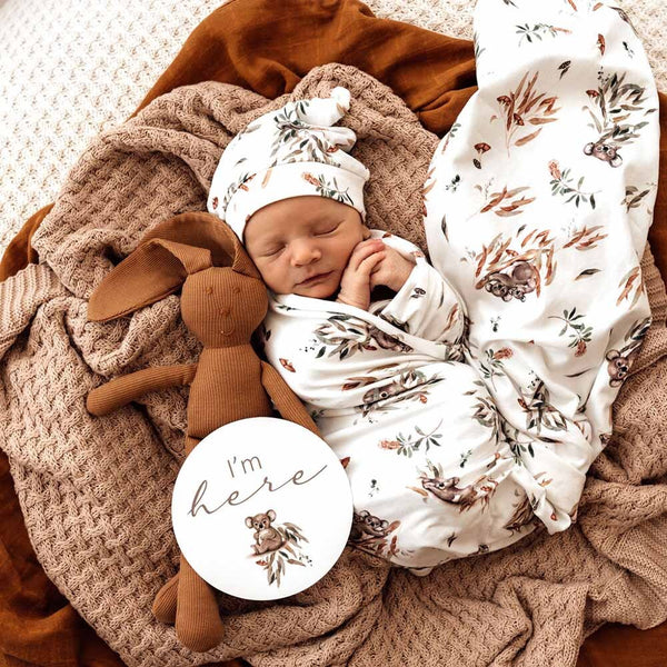 Newborn baby wrapped in a floral blanket with a brown plush toy, surrounded by soft textures.