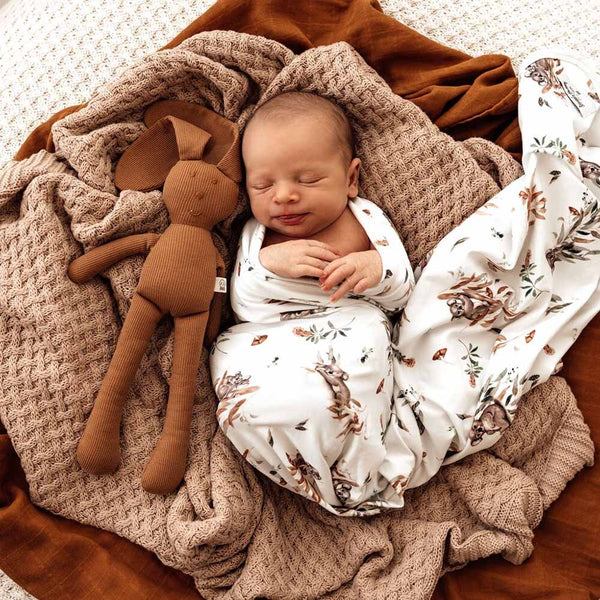 Newborn baby wrapped in a patterned blanket with a brown stuffed toy on a wooden floor.