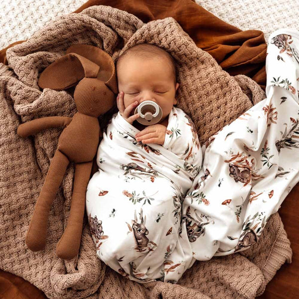 Newborn baby wrapped in a blanket with animal patterns, holding a pacifier, next to a brown stuffed toy on a soft surface.