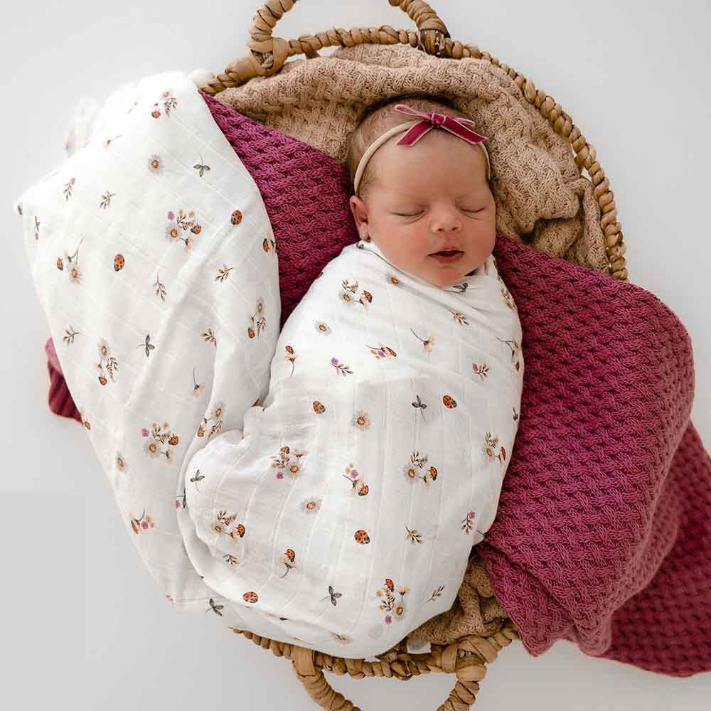 Newborn baby swaddled in a floral blanket with a pink headband, lying on a textured blanket in a woven basket.