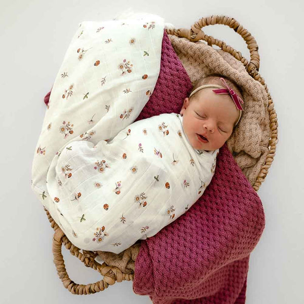 Newborn baby swaddled in a floral blanket with a pink headband, lying on a textured blanket in a wicker basket.