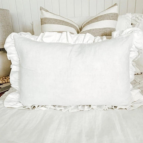 White pillow with ruffled edges on a bed with neutral bedding and lamp in the background.