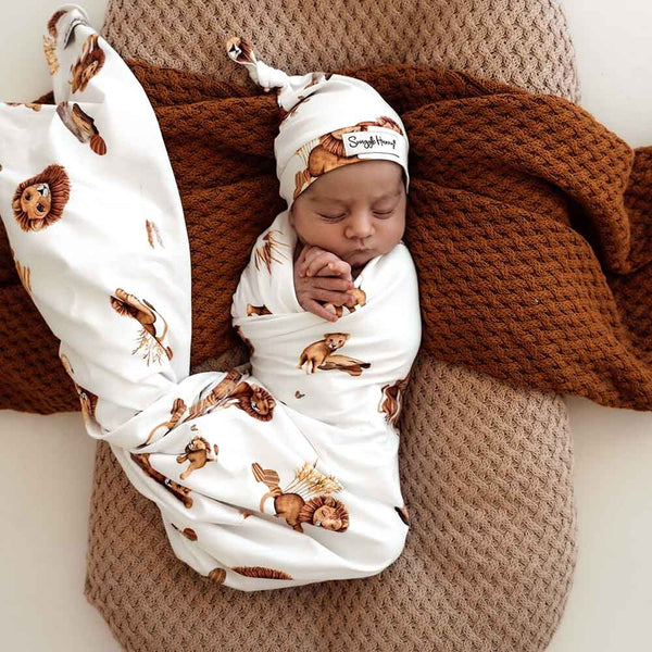 Newborn baby wrapped in a lion-patterned swaddle with a matching hat, lying on a brown textured blanket.