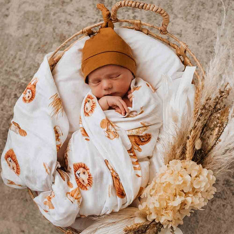 Newborn baby wrapped in a lion-patterned blanket, wearing a brown hat, in a woven basket with flowers.