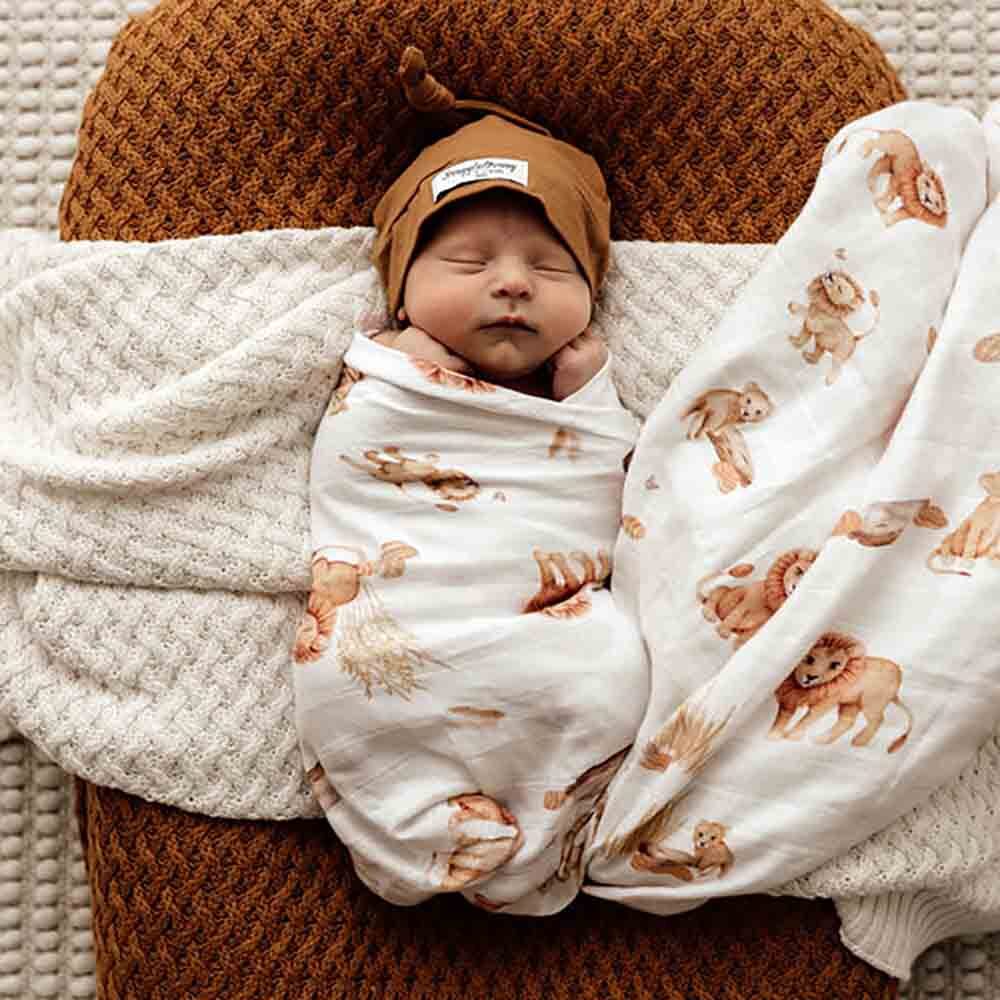 Newborn baby swaddled in a blanket with lion patterns, wearing a brown hat.
