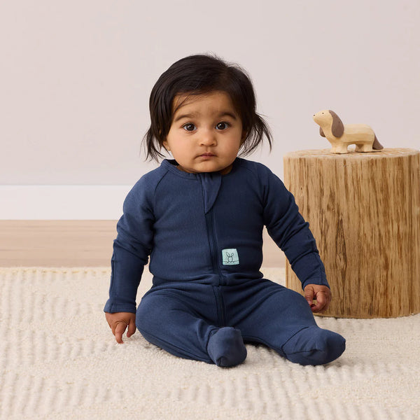 Baby wearing a navy blue onesie sitting on a beige carpet with a wooden stool and toy in the background.