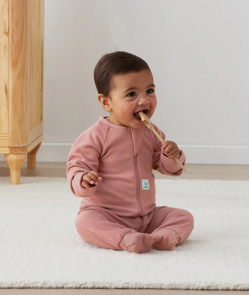 Baby in pink outfit sitting on a white rug with a wooden cabinet in the background
