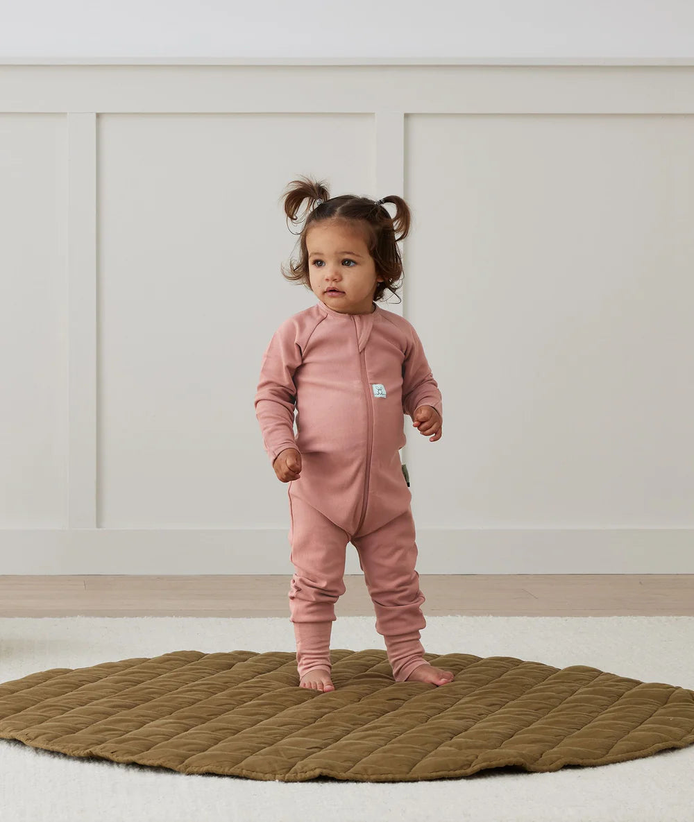 Child wearing a pink onesie standing on a textured rug with a neutral background
