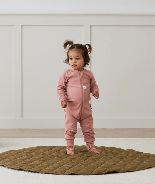 Child wearing a pink onesie standing on a textured rug with a neutral background