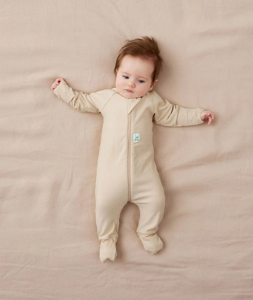 Baby lying on a beige blanket wearing a beige onesie with a small logo on the chest.