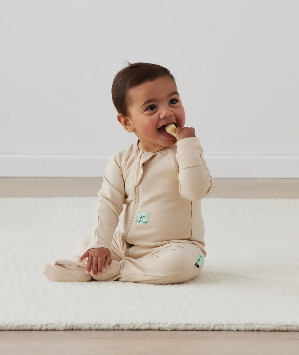 Baby in a beige outfit sitting on a white surface, eating a snack.