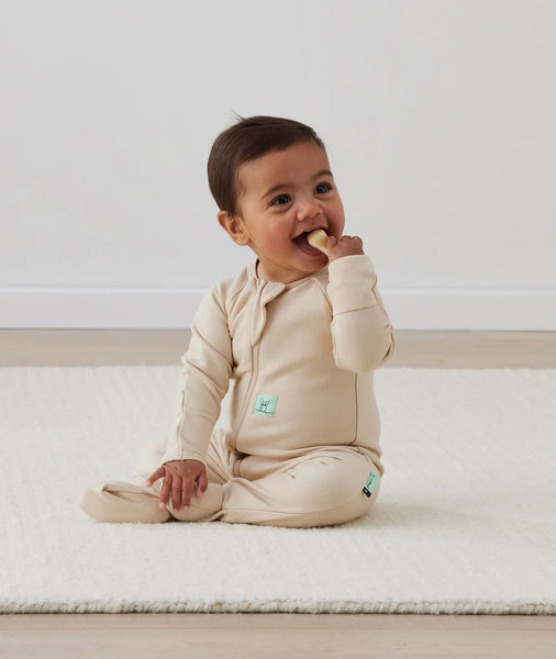 Baby in a beige outfit sitting on a white surface, eating a snack.
